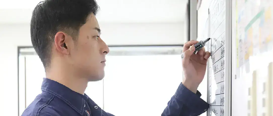 作業服を着た男性がホワイトボードにマーカーで予定を書き込んでいる様子。